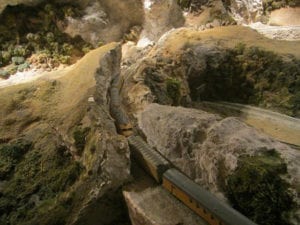 The model train passes through a narrow passage between two rocky mountain walls.