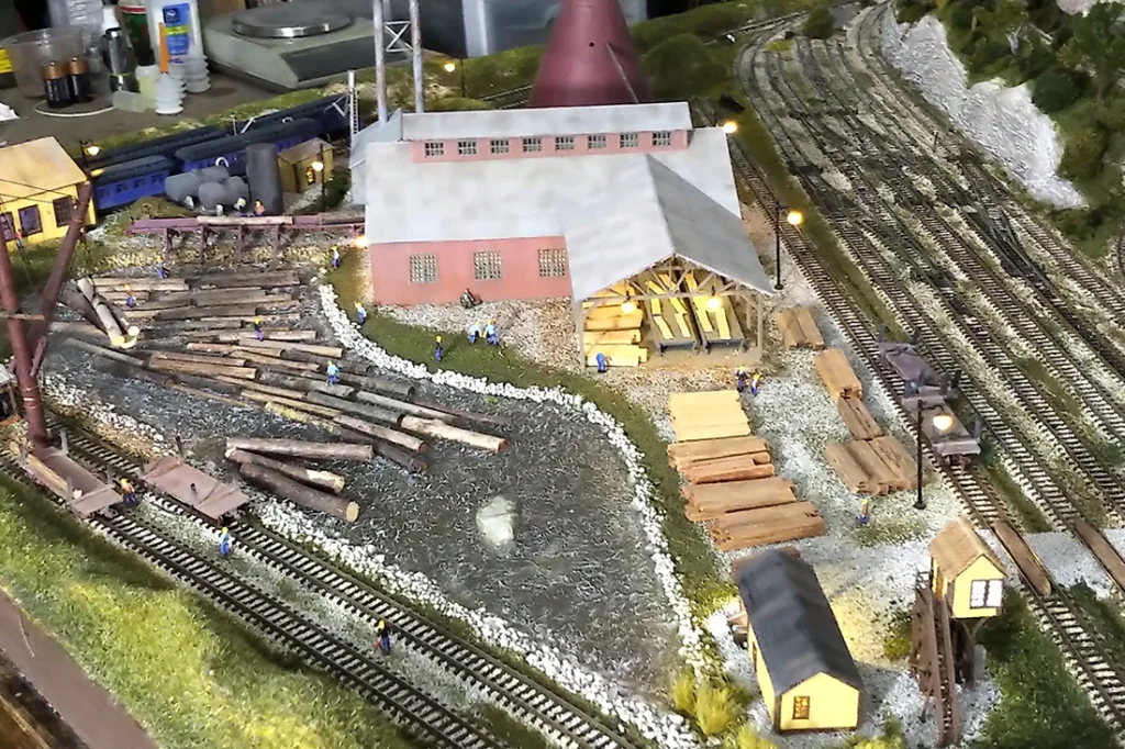 HO scale model train layout featuring a detailed logging railroad scene with a bustling lumber mill, workers, and logs.