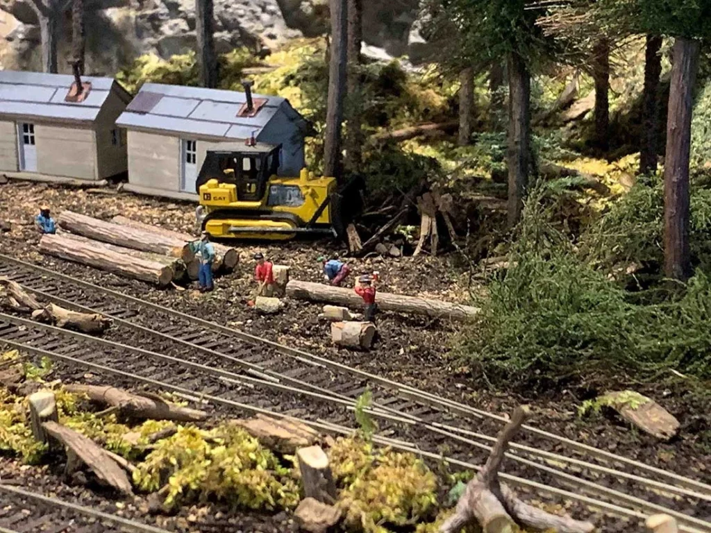 logging scene with a yellow bulldozer, small figures, logs, and two gray cabins amidst trees and train tracks