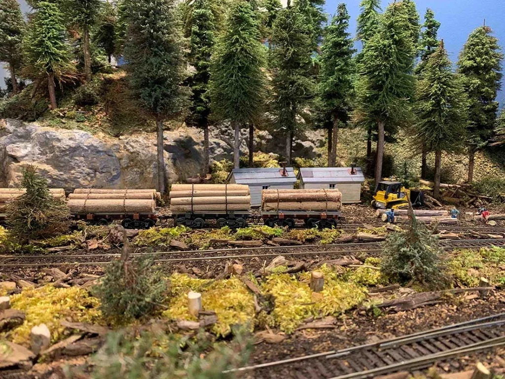 model railroad depicting a logging scene with trees, a train carrying logs, small buildings, and a construction vehicle in a forested area.