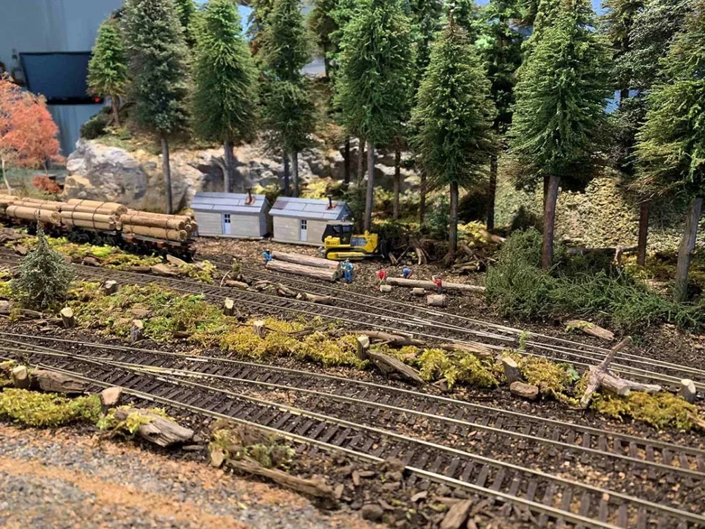 model railroad with logging scene, featuring tracks, logs, trees, small buildings, and workers in a forest setting