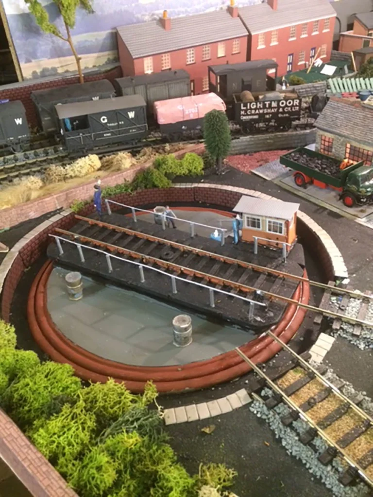 O gauge train layout with turntable, water tower, goods yard, and detailed scenery including buildings and trees.